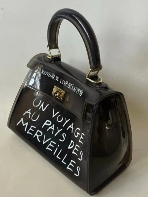 Svart transparent handväska med text - Cool svart transparent handväska i plast med vit text 'UN VOYAGE AU PAYS DES MERVEILLES' och guldfärgade metalldetaljer. Väskan har ett topphandtag och axelrem, samt vridlås framtill. Perfekt statement piece för dig som gillar unik stil.