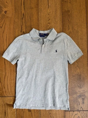  pikétröja från Polo Ralph Lauren 14-16 år - Klassisk ljusgrå pikétröja från Polo Ralph Lauren med korta ärmar och krage. Tröjan har två knappar vid halsen och den ikoniska lilla loggan broderad på bröstet. Perfekt till jeans eller shorts för en clean och enkel stil.