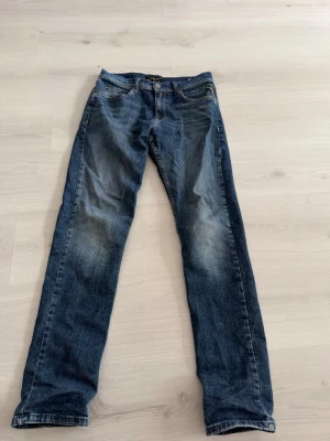 Blå jeans från Replay - Snygga blå jeans från Replay med klassisk femficksmodell och raka ben. Jeansen har en mörk tvätt med ljusa slitningar framtill och detaljerade sömmar. Perfekta för dig som gillar en tidlös och avslappnad stil.
