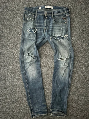 Blå slitna jeans från Replay - Säljer ett par blå jeans från Replay med skinny fit och flera slitningar och hål på både fram- och baksida. Jeansen har klassisk femficksdesign, låg midja och detaljerade slitningar för en trendig look. Perfekta för dig som gillar en edgy stil. Jeansen har även blivit lagade i skrevet men det är knappast synligt.