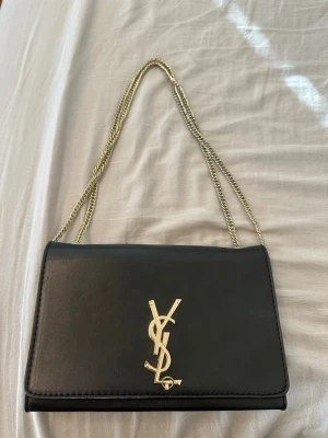 Svart axelväska med guldkedja YSL - Snygg svart axelväska från Yves Saint Laurent med guldfärgad kedjerem och ikonisk YSL-logga framtill. Väskan har en stilren, rektangulär form och är tillverkad i slätt läder. Perfekt för dig som gillar klassisk och lyxig design.