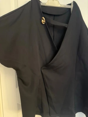 Svart omlottopp med knappdetalj - Snygg svart omlottopp med unik guldknapp vid axeln. Toppen har lös passform och korta ärmar, vilket ger en cool och avslappnad look. Perfekt för dig som gillar stilrena plagg med en twist.