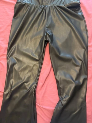 Svarta utsvängda leggings i fuskskinn - Snygga svarta leggings i glansigt fuskskinn med utsvängda ben. De har hög midja och resår i midjan för skön passform. Perfekta för dig som vill ha en edgy och trendig look. Materialet är mjukt och följsamt.