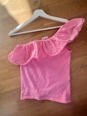 Rosa offshoulder topp från Kappahl - Söt rosa offshoulder topp från Kappahl med volangdetalj längs axeln. Toppen är ärmlös och har en asymmetrisk design som ger en trendig look. Tillverkad i mjuk bomull, perfekt för varma dagar.
