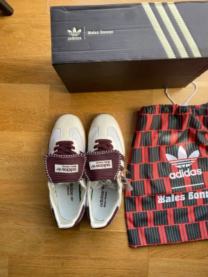 Adidas Wales Bonner sneakers vita/vinröda - Unika Adidas Wales Bonner sneakers i vitt och vinrött med snygga kontrastsömmar och klassisk retrostil. Skorna har lågt skaft, ovandel i mocka och textil samt detaljerad plös med Wales Bonner-logga. Kommer med originalkartong och dustbag.