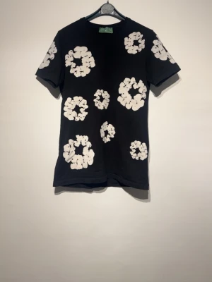 Svart t-shirt med vita blommor - Snygg svart t-shirt med stora vita blommönster över hela tröjan. Klassisk passform och korta ärmar, perfekt för dig som vill sticka ut lite extra. Tillverkad i mjuk bomull som känns skön mot huden.