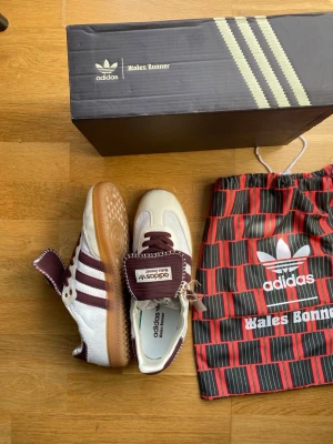 Adidas Wales Bonner sneakers vita/vinröda - Snygga Adidas Wales Bonner sneakers i vitt och vinrött med klassiska tre ränder på sidan och gummisula. Skorna har unika detaljer som grova sömmar och bred plös med Wales Bonner-logga. Materialmix av läder och textil. Kommer med originalkartong och dustbag.