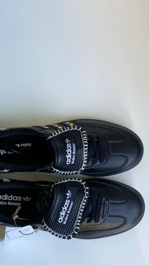Adidas Wales Bonner svarta loafers - Svarta loafers från Adidas x Wales Bonner med vita kontrastsömmar och gulddetaljer. Skorna har en klassisk siluett med snörning och branding på plösen och innersulan. Tillverkade i läder med unika detaljer som ger en cool och modern vibe.