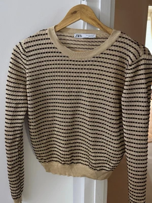 Beige stickad tröja från Zara - Snygg beige stickad tröja från Zara med svarta horisontella ränder och rund halsringning. Tröjan har ribbade muddar vid ärmslut och nederkant, och ett coolt rutmönster som ger en modern vibe.