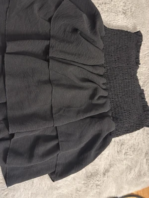 Svart volangkjol med smock - Kort svart kjol med bred smock i midjan och flera lager volanger. Kjolen har en luftig och trendig look, perfekt för att styla med en crop top eller stickad tröja. Materialet känns mjukt och följsamt.