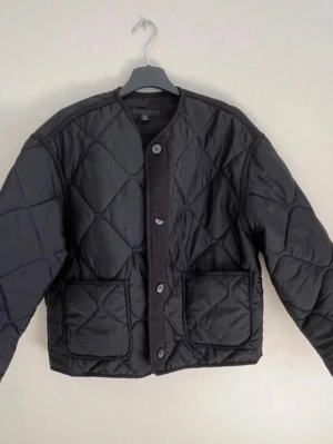 Svart quiltad pufferjacka från ZARA - Stilren svart pufferjacka med stora diamantformade quiltade sömmar. Jackan har rund halsringning, knäppning framtill med knappar och två stora fickor fram. Materialet är glansigt och ger en modern look. Perfekt för kalla dagar och en knapp blivit halva 👌