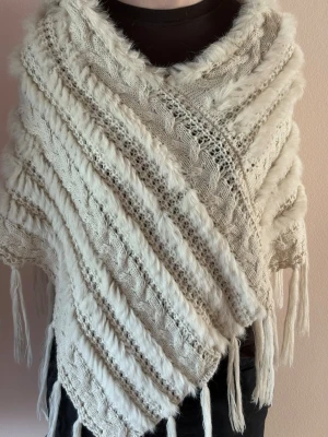 Beige stickad poncho med luva - Säljer en mysig beige stickad poncho med luva och fransar. Ponchon har ett fint kabelstickat mönster och inslag av fluffiga detaljer som ger en unik look. Perfekt att slänga över en tröja för extra värme och stil. Str One size