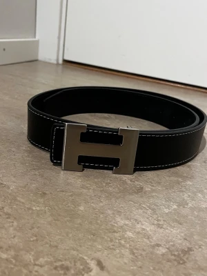 Svart skärp med stor metallspänne - Snyggt svart bälte i skinn med ett stort, silverfärgat metallspänne i form av ett H. Bältet har vita kontrastsömmar längs kanterna som ger en stilren look. Perfekt accessoar för att lyfta din outfit.