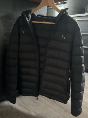 Svart dunjacka från Polo Ralph Lauren - Snygg svart dunjacka från Polo Ralph Lauren med huva och dragkedja framtill. Jackan har det klassiska Polo-logotypen broderad på bröstet och quiltad design som ger en modern look. Perfekt nu till hösten då den värmer men inte är för varm. Priset är ej hugget i sten men de låga priset är för ena dragkedjan saknar själva grejen du drar i. BUDA