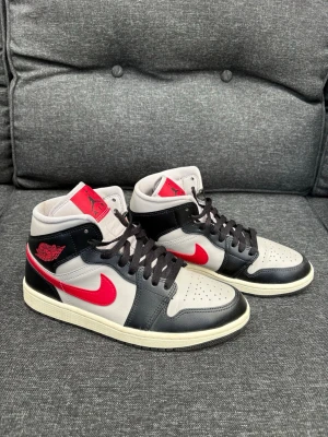 Nike Air Jordan 1 Mid svart/röd/grå - Snygga Nike Air Jordan 1 Mid sneakers i svart, grått och rött. Skorna har klassisk röd Swoosh, svarta läderpaneler och grå bas. Röd logga på sidan och tunga. Tjock vit sula och svarta skosnören. Perfekta för dig som gillar streetwear och ikonisk design.