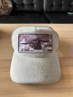 Ljusgrå keps med Mbappé - Ljusgrå keps i mockaliknande material med mesh baktill och justerbar snapback. Framtill finns ett fototryck av Mbappé. Märkt med Evosportswear på insidan. Perfekt för dig som gillar sportig stil.