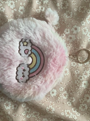 Fluffig rosa plånbok med regnbåge - Supersöt rund plånbok i fluffigt rosa material med broderad regnbåge och moln på framsidan. Plånboken har dragkedja och en liten tofs samt nyckelring, perfekt att hänga på väskan. Insidan är fodrad i ljusrosa tyg.