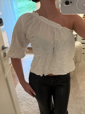 Vit oneshoulder topp med puffärm - Säljer en vit oneshoulder topp med puffärm och resår i halsringning. Toppen är croppad och har en luftig passform, perfekt för dig som vill ha en unik och trendig look. 