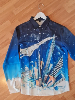 Rave Party Outfit - Unik långärmad skjorta från Casablanca med ett färgstarkt tryck av stadssilhuetter, stjärnhimmel och Concorde-flygplan. Skjortan har knappar framtill och en klassisk krage. Materialet känns glansigt och lyxigt, perfekt för dig som vill sticka ut.
