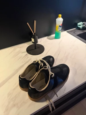 maision margiela - Säljer ett par stilrena svarta sneakers med beige detaljer och gummisula. Skorna har vita snören och en klassisk låg profil. Materialet ser ut att vara en mix av läder och mocka, vilket ger en snygg kontrast. Perfekta för dig som gillar minimalistisk och tidlös stil.