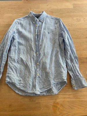 Blårutig skjorta ralph lauren skjorta - Klassisk blårutig skjorta med button-down krage och långa ärmar. Skjortan har vita och blå rutor, knappar framtill och vid ärmsluten samt en liten broderad logga på bröstet. Tillverkad i bomull för en skön och luftig känsla.