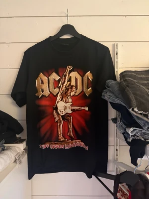 Svart AC/DC T-shirt med tryck - Säljer en svart AC/DC T-shirt med stort tryck av en gitarrist och texten 'Stiff Upper Lip Tour' på framsidan. T-shirten har klassisk passform och korta ärmar. Perfekt för dig som gillar rock och vill sticka ut med en snygg bandtröja.