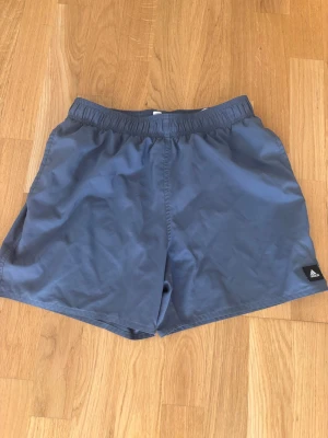 Blå badshorts från Adidas - Säljer ett par blå badshorts från Adidas med elastisk midja och snörning. Shortsen har meshfoder på insidan och en liten Adidas-logga på benet. 