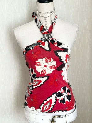 Röd blommig halterneck topp - Snygg halterneck topp med blommigt mönster i rött, svart och vitt. Toppen har en omlott-detalj vid halsen och är ärmlös med figurnära passform. Perfekt för dig som vill sticka ut med färg och mönster.