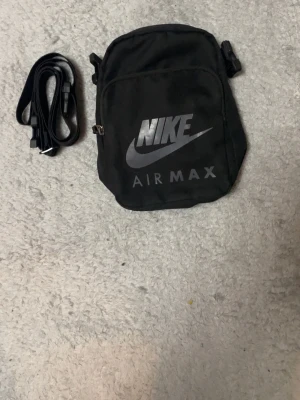 Svart Nike Air Max axelväska ingår också bandet till väskan - Snygg svart axelväska från Nike Air Max med justerbar rem och stort logotryck framtill. Väskan har dragkedja och är tillverkad i slitstarkt tygmaterial. Perfekt för att bära det viktigaste och passar till streetwear-stilen.