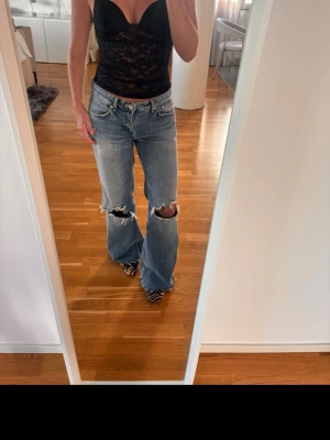 Ljusblå slitna jeans - Säljer ett par ljusblå bootcut jeans med stora slitningar vid knäna och råa kanter. Jeansen har klassisk femficksdesign och låg midja. Perfekta för en avslappnad och trendig look.