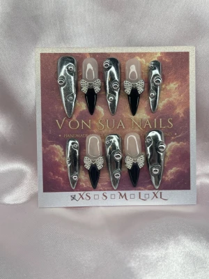 BadAngel (XS);Medium Stiletto - Unika handgjorda press-on naglar i silver och svart. Naglarna har en medium lång stiletto form och dekoreras med 3D-detaljer, svart french och silvriga rosetter. Designen är handgjord för en edgy och konstnärlig look.XS: 14 mm, 11 mm, 11,5 mm, 10 mm, 8 mm