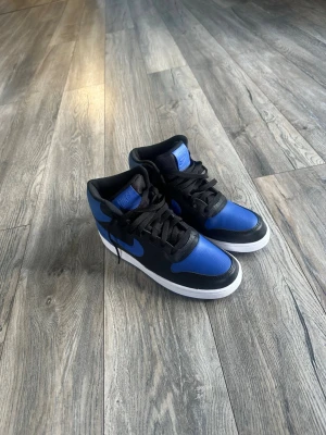 Nike blå och svarta high sneakers - Snygga Nike sneakers i high-modell med blå och svarta paneler, vita sulor och klassisk Nike swoosh på sidan. Ovandelen är i textil och syntet, med matchande blå detaljer på plös och yttersula. Perfekta för dig som vill sticka ut med stilrena färger.