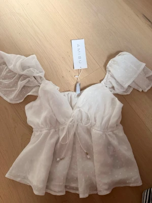 Vit blus med volangärmar från Amisu - Söt vit blus från Amisu med transparenta volangärmar och knyt i midjan. Blusen har ett diskret prickigt mönster och v-ringad hals. Perfekt för sommarens alla tillfällen och ger en romantisk vibe. Storlek M men passar S