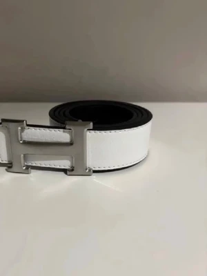Vitt skärp med silverfärgat H-spänne - Snyggt vitt skärp med ett stort H-format spänne i silverfärg. Bältet har stilrena sömmar och är gjort i slätt skinn. Perfekt accessoar för att lyfta din outfit.