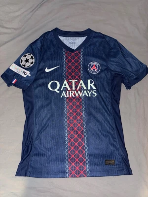 PSG matchtröja Nike blå röd - Snygg PSG matchtröja i mörkblått med röd och vit detalj på framsidan. Tröjan har Qatar Airways som sponsor, Champions League-märke på ärmen och spelarnamn D. Doué samt nummer 14 på ryggen. Tillverkad i lätt och ventilerande material.