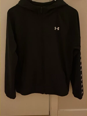 Svart vindjacka från Under Armour - Snygg svart vindjacka från Under Armour med dragkedja framtill och huva. Logga på bröstet och text på ena ärmen ger en sportig vibe. Tillverkad i ett mjukt och bekvämt material, perfekt för chill eller träning.