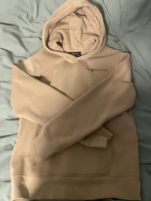 Beige hoodie från Polo Ralph Lauren - Säljer en beige hoodie från Polo Ralph Lauren med klassisk huva och stor magficka. Diskret logga broderad på bröstet. Hoodien har en avslappnad passform och är gjord i mjukt material som känns skönt mot huden. Perfekt för en chill och stilren look.