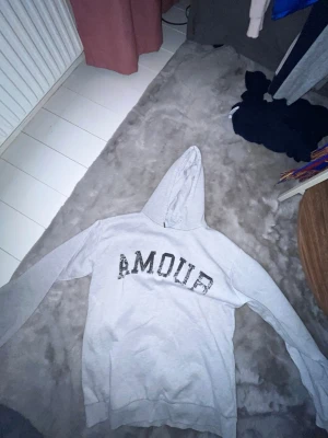 Ljusgrå hoodie med tryck AMOUR - Säljer en ljusgrå hoodie med huva och texten AMOUR i svart glittertryck på bröstet. På baksidan finns en stor stjärna i samma stil. Hoodien har ribbade muddar vid ärmslut och nederkant. Perfekt för en chill och trendig look.