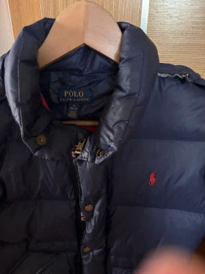 Mörkblå dunjacka från Polo Ralph Lauren strlk M - 10-12 år - Snygg mörkblå dunjacka från Polo Ralph Lauren  med hög krage, dragkedja och knappar framtill. Jackan har två stora fickor med dragkedja och tryckknappar samt den klassiska röda loggan broderad på bröstet. Perfekt för kalla dagar. Stlrl M, 10-12 år