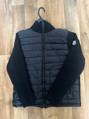 Svart cardigan - Snygg svart cardiganjacka från  med quiltad front och rygg i blankt material samt stickade ärmar och krage. Dragkedja framtill och logga på ärmen. Perfekt för lager-på-lager och streetwear-vibe.