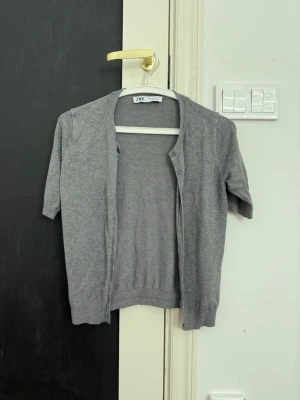 Grå kortärmad kofta från ZARA - Stilren grå kofta från ZARA med korta ärmar och knäppning framtill. Tillverkad i mjukt, tunt material som passar perfekt för lager på lager. Enkel och clean design som funkar till det mesta.