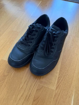 Svarta sneakers med snörning - Säljer ett par helsvarta sneakers med snörning och perforerade detaljer på tån och sidan. Skorna har en klassisk låg siluett och är tillverkade i syntetmaterial med gummisula. Perfekta för en clean och stilren look.