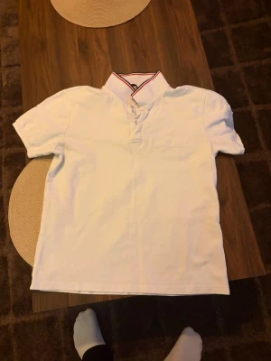 Vit Moncler piké med röd detalj - Snygg vit pikétröja från Moncler med korta ärmar och klassisk krage med röd och marinblå rand. Broderad Moncler-logga på ärmen. Perfekt för en stilren och sportig look.
