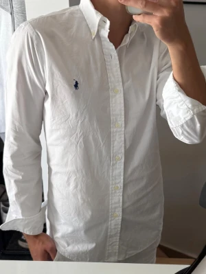 Vit skjorta från Ralph Lauren - Klassisk vit skjorta från Ralph Lauren med blå broderad logga på bröstet. Skjortan har lång ärm, knappar framtill och krage. Tillverkad i bomull för en skön och stilren känsla. Perfekt till många olika looks.