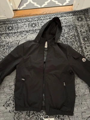 Svart vindjacka med huva från Moncler - Snygg svart vindjacka från Moncler med huva och dragkedja framtill. Jackan har två fickor med dragkedja och en ficka med knapp på ärmen där Moncler-loggan sitter. Tillverkad i ett lätt och vattenavvisande material, perfekt för blåsiga dagar.