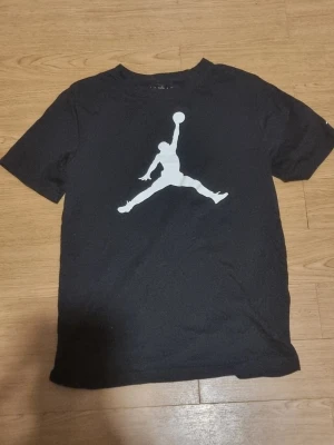 Svart Jordan t-shirt med stor logga - Snygg svart Jordan t-shirt med en stor vit logga på bröstet. Klassisk rund hals och korta ärmar. Tillverkad i mjukt bomullsmaterial som är skönt att bära. Perfekt för dig som gillar sportig och stilren design.