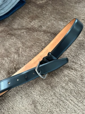 Svart klassiskt skärp i skinn - Snyggt svart bälte i äkta skinn med enkel silverfärgad metallspänne. Bältet har en stilren och tidlös design som passar till många olika outfits. Insidan är ljust beige och bältet har flera hål för justerbar passform.