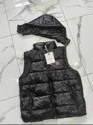 Svart dunväst från Moncler - Snygg svart dunväst från Moncler med avtagbar huva och dragkedja framtill. Västen har två fickor med dragkedja och Moncler-logga på bröstet. Materialet är glansigt och quiltat för en modern look. Fins i alla storlekar ⚠️