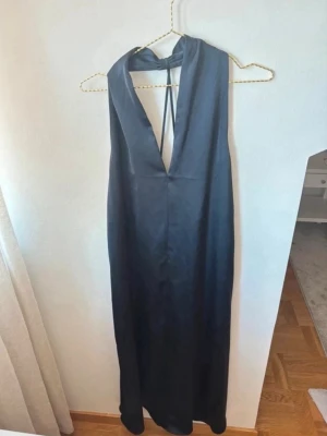 Marinblå långklänning med öppen rygg - Stilren marinblå långklänning i ett glansigt material med djup v-ringning och öppen rygg. Klänningen har halterneck med knyt i nacken och ett elegant fall. Perfekt för dig som vill ha en enkel men snygg look.