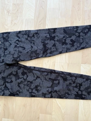 Mörkgrå leggings med camouflage-mönster - Säljer ett par mörkgrå leggings med svart camouflage-mönster. De har en tight passform och är perfekta för träning eller chill. Materialet känns mjukt och stretchigt, vilket gör dem bekväma att bära hela dagen.